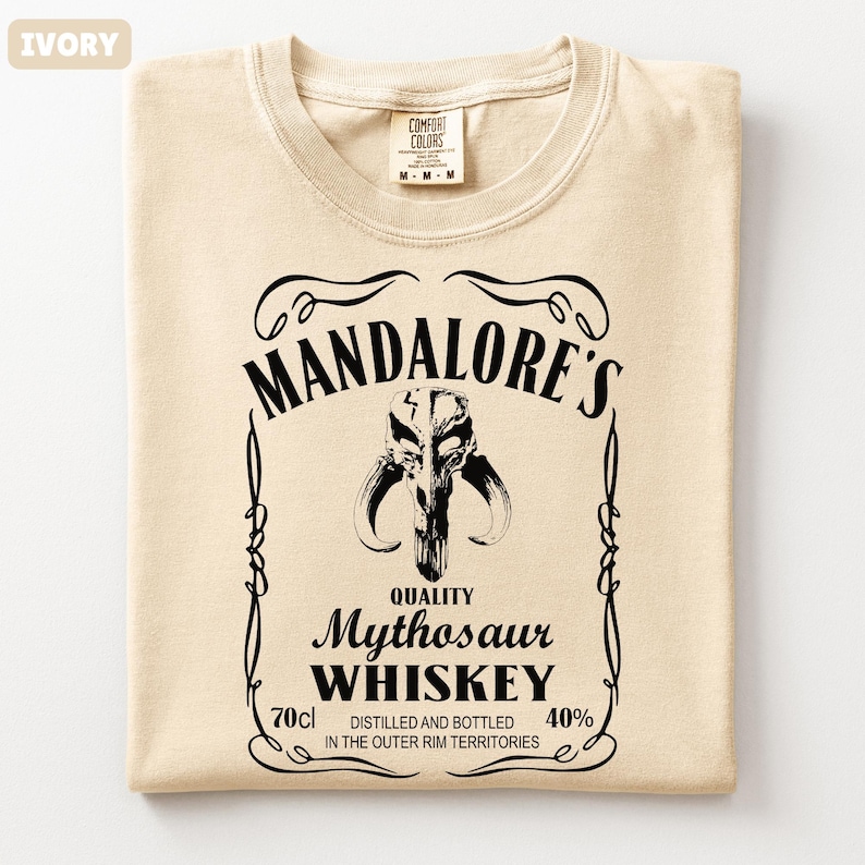 Puede incluir: Camiseta de color marfil con un gr&aacute;fico negro que se asemeja a una etiqueta de whisky. La etiqueta dice "MANDALORE'S Mythosaur WHISKEY" con un emblema de calavera. La camiseta tambi&eacute;n tiene el texto "70cl" y "40%" en la etiqueta.