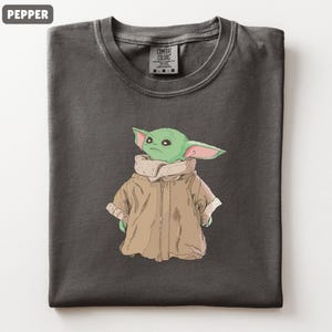May include: A dark gray t-shirt featuring a cartoon illustration of a green alien character with large ears and a tan coat. The shirt has a crew neck and a tag that reads "Comfort Colors".