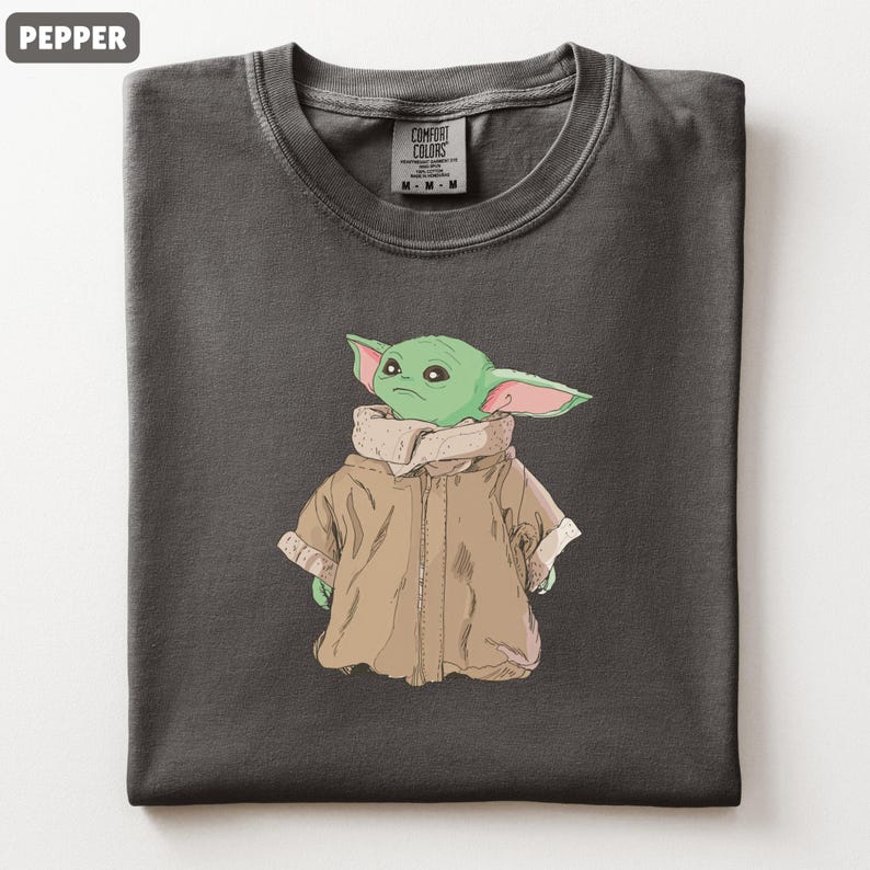 May include: A dark gray t-shirt featuring a cartoon illustration of a green alien character with large ears and a tan coat. The shirt has a crew neck and a tag that reads "Comfort Colors".