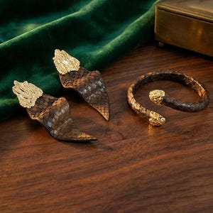 Puede incluir: Un conjunto de joyas con un par de pendientes y una pulsera. Los pendientes y la pulsera tienen un patrón de escamas de serpiente en tonos marrones y dorados. Los pendientes y la pulsera tienen detalles dorados.