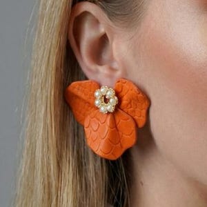 Op de afbeelding: Oranje bloemvormige oorbellen met een getextureerd oppervlak en een cluster van kleine, ronde, witte parels in het midden. De oorbellen worden gedragen door een persoon met blond haar en een wit overhemd met kraag.