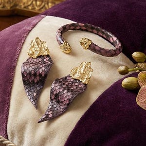 May include: A jewellery set comprising a bracelet and earrings with a snakeskin pattern in purple and pink hues. The earrings and bracelet have gold-coloured accents. The items are displayed on a purple, cream, and lavender cushion.