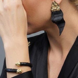 Puede incluir: Pendientes de cuero negro con detalles dorados. Los pendientes tienen una forma abstracta &uacute;nica. Tambi&eacute;n se ve una pulsera de cuero negro a juego con detalles dorados. Las joyas se llevan con una parte superior negra.