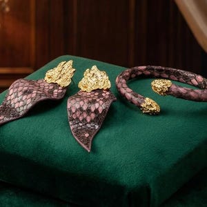 Puede incluir: Un conjunto de joyas con pendientes y una pulsera con un patr&oacute;n de escamas de serpiente en rosa y marr&oacute;n. Los pendientes y la pulsera tienen detalles dorados. El conjunto se exhibe sobre una superficie de terciopelo verde, con dos cajas doradas ornamentadas en el fondo.