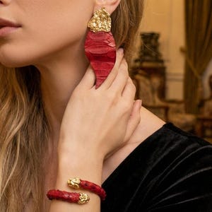 May include: Red and gold-toned jewelry set. The set includes a pair of large, teardrop-shaped earrings and a matching cuff bracelet. The earrings and bracelet feature a textured red material with gold-colored accents.
