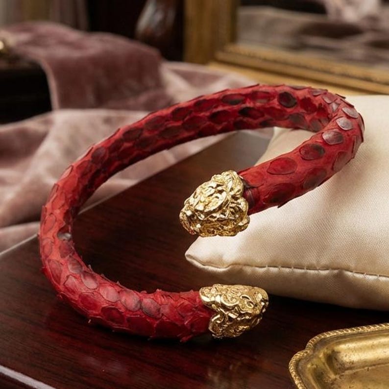 Puede incluir: Una pulsera de piel de serpiente roja con detalles de cabeza de le&oacute;n dorada. La pulsera abierta descansa sobre un coj&iacute;n blanco. Un accesorio de moda llamativo.