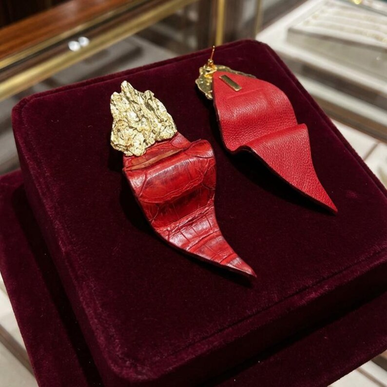 Puede incluir: Un par de pendientes llamativos. Un pendiente presenta una parte superior dorada texturizada y una forma de cuerno de cuero rojo con textura de cocodrilo. El otro pendiente tiene una parte superior dorada y un dise&ntilde;o de cuero rojo plegado. Los pendientes se exhiben sobre terciopelo burdeos.