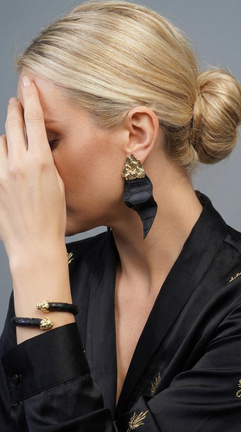Puede incluir: Pendientes de cuero negro con detalles dorados. Los pendientes tienen una forma abstracta &uacute;nica. Tambi&eacute;n se ve una pulsera de cuero negro a juego con detalles dorados. Las joyas se llevan con una parte superior negra.