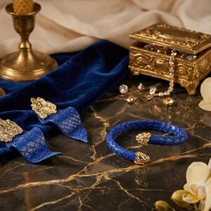 Puede incluir: Una elegante exhibici&oacute;n con una pulsera y pendientes de cuero azul con detalles dorados. Una caja de joyer&iacute;a dorada, velas y orqu&iacute;deas completan la est&eacute;tica lujosa. Los art&iacute;culos est&aacute;n dispuestos sobre una superficie de m&aacute;rmol.