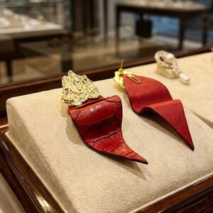 Puede incluir: Un par de pendientes de cuero rojo con detalles dorados. Los pendientes tienen un dise&ntilde;o texturizado y plegado, con una pieza dorada en la parte superior. Los pendientes se exhiben sobre una superficie beige.