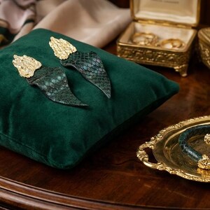 Puede incluir: Pendientes y pulsera con detalles dorados y un dise&ntilde;o de piel de serpiente verde. Los pendientes est&aacute;n sobre un coj&iacute;n de terciopelo verde. La pulsera est&aacute; en una bandeja dorada.