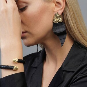 Puede incluir: Pulsera de cuero negro con detalles dorados. Se ve un pendiente negro grande con la parte superior dorada. La imagen muestra a una persona que lleva las joyas. El fondo es gris liso.