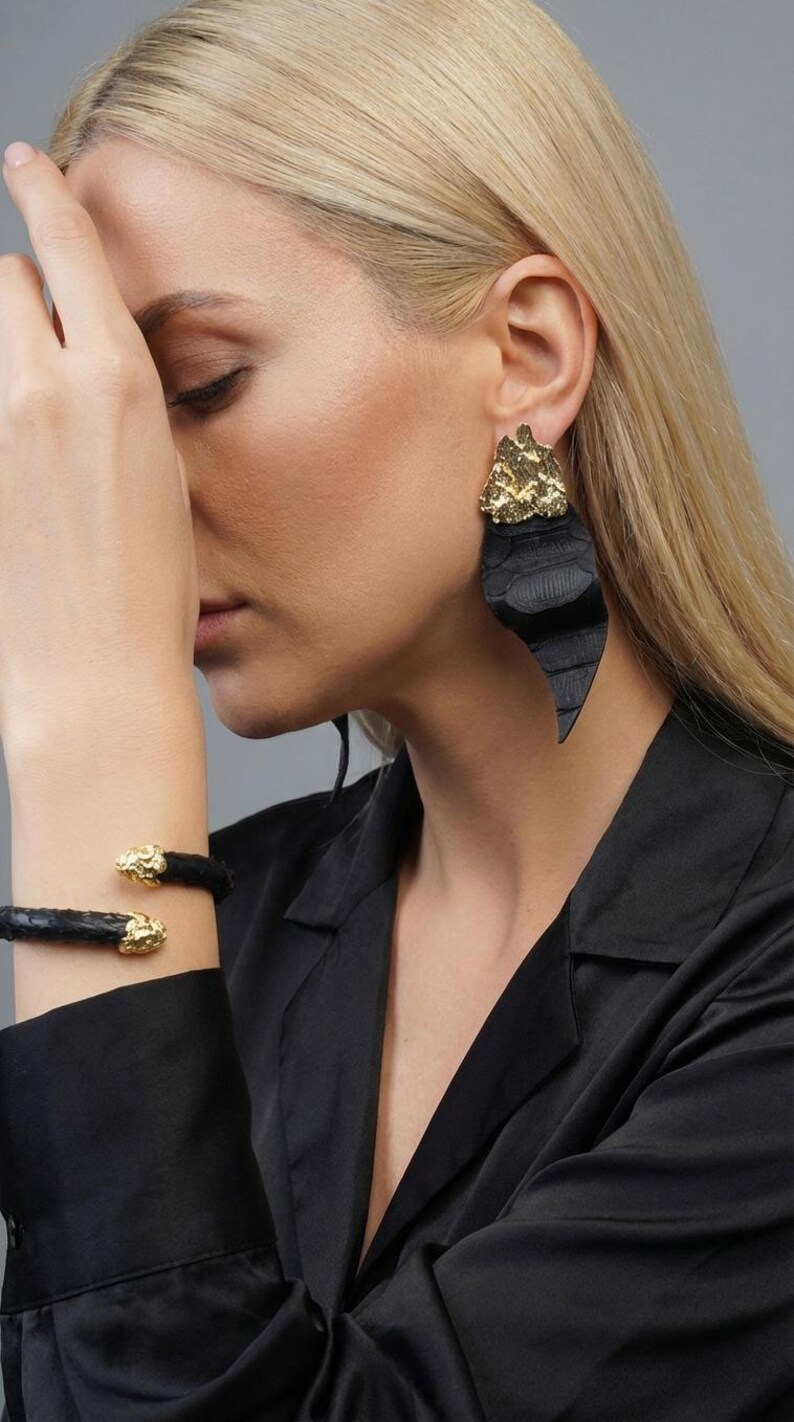 Puede incluir: Pulsera de cuero negro con detalles dorados. Se ve un pendiente negro grande con la parte superior dorada. La imagen muestra a una persona que lleva las joyas. El fondo es gris liso.