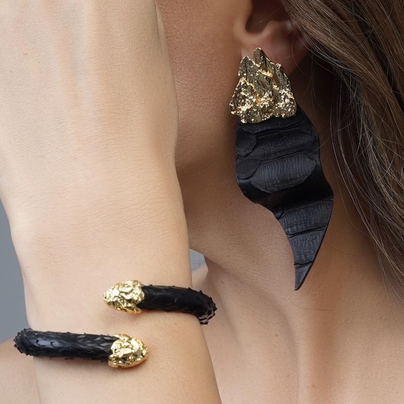 Puede incluir: Pulsera de pu&ntilde;o y pendientes de cuero negro con detalles dorados. Los pendientes tienen una parte superior dorada texturizada y una parte inferior larga y curvada de cuero negro. La pulsera de pu&ntilde;o tiene un dise&ntilde;o similar.