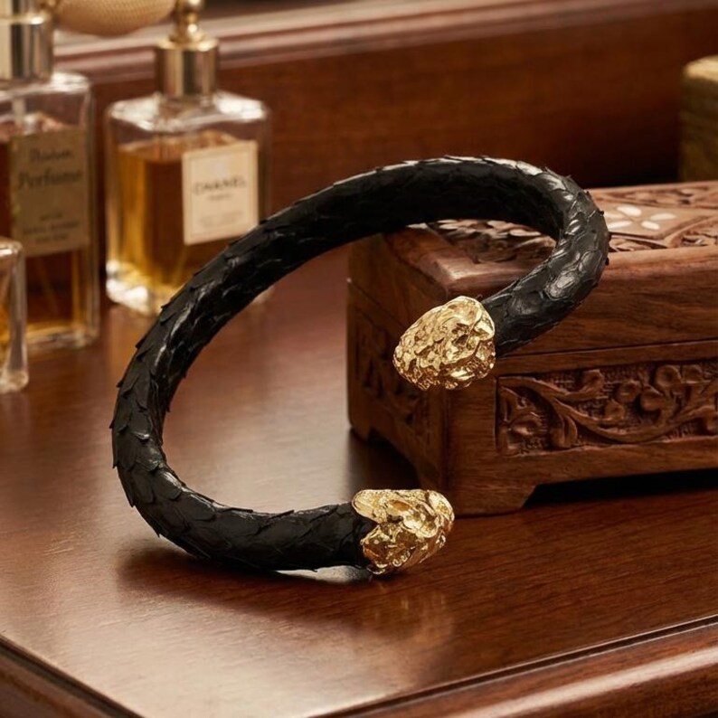 Puede incluir: Una pulsera de cuero negro con extremos texturizados de color dorado. La pulsera abierta descansa sobre una superficie de madera, con frascos de perfume y una caja decorativa en el fondo. La pulsera tiene una textura similar a la de una serpiente.
