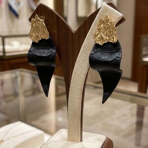 Puede incluir: Un par de pendientes llamativos en exhibici&oacute;n. Cada pendiente presenta un dise&ntilde;o de cuero negro texturizado con una parte superior dorada irregular. Los pendientes se exhiben en un soporte de madera con una tela color crema.