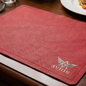 May include: A red leather placemat with a textured surface. The placemat has a stitched border and a logo that says "AVIOR" in silver and white. Silverware and a plate of food are in the background.