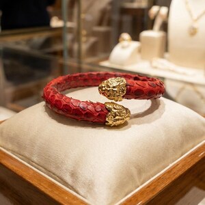 Puede incluir: Una pulsera de piel de serpiente roja con detalles de cabeza de le&oacute;n dorada. La pulsera se exhibe sobre un coj&iacute;n crema dentro de una vitrina de madera. La joyer&iacute;a est&aacute; en un entorno de tienda.