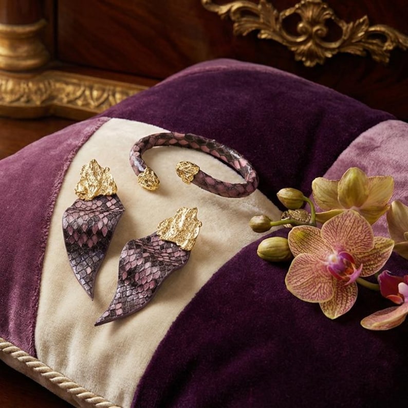 Puede incluir: Un conjunto de joyas con una pulsera y pendientes con un estampado de piel de serpiente en tonos morados y rosas. Los pendientes y la pulsera tienen detalles dorados. Los art&iacute;culos se exhiben sobre un coj&iacute;n morado, crema y lavanda.
