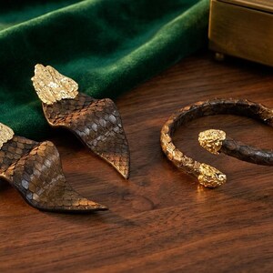 Puede incluir: Un conjunto de joyas con pendientes y una pulsera. Los pendientes y la pulsera est&aacute;n hechos de materiales marrones y dorados, con un dise&ntilde;o de serpiente. Los pendientes tienen una parte superior dorada, y la pulsera tiene cabezas de serpiente doradas en cada extremo.