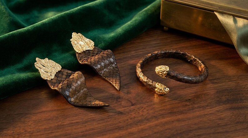 Puede incluir: Un conjunto de joyas con pendientes y una pulsera. Los pendientes y la pulsera est&aacute;n hechos de materiales marrones y dorados, con un dise&ntilde;o de serpiente. Los pendientes tienen una parte superior dorada, y la pulsera tiene cabezas de serpiente doradas en cada extremo.