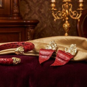 Puede incluir: Un brazalete de piel de serpiente roja y pendientes a juego con detalles dorados. Las joyas se exhiben sobre una superficie de terciopelo burdeos con una tela dorada drapeada. El fondo incluye muebles antiguos y un candelabro.
