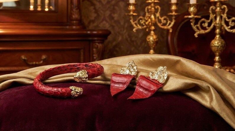 Puede incluir: Un brazalete de piel de serpiente roja y pendientes a juego con detalles dorados. Las joyas se exhiben sobre una superficie de terciopelo burdeos con una tela dorada drapeada. El fondo incluye muebles antiguos y un candelabro.