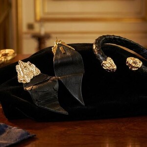 Puede incluir: Una colecci&oacute;n de joyas de lujo exhibidas sobre una superficie de terciopelo negro. El conjunto incluye una pulsera de cuero negro con detalles dorados y un par de pendientes de cuero negro con detalles dorados. Otros objetos decorativos dorados est&aacute;n en el fondo.