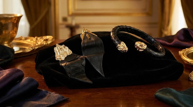 Puede incluir: Una colecci&oacute;n de joyas de lujo exhibidas sobre una superficie de terciopelo negro. El conjunto incluye una pulsera de cuero negro con detalles dorados y un par de pendientes de cuero negro con detalles dorados. Otros objetos decorativos dorados est&aacute;n en el fondo.