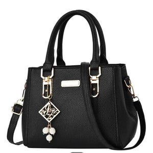 May include: Black handbag with top handles and a detachable shoulder strap. The bag features gold-tone hardware, a decorative charm, and a textured exterior. The bag has a rectangular shape with a structured design.