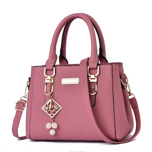 May include: A dusty rose handbag with a structured design. It features top handles, a detachable shoulder strap, and gold-tone hardware. A decorative charm with a diamond-shaped pendant and pearl accents hangs from the front.