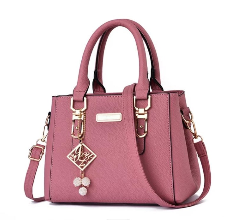 May include: A dusty rose handbag with a structured design. It features top handles, a detachable shoulder strap, and gold-tone hardware. A decorative charm with a diamond-shaped pendant and pearl accents hangs from the front.