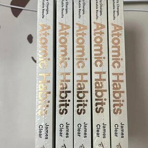 Peut inclure: Cinq exemplaires du livre "Atomic Habits" de James Clear sont pr&eacute;sent&eacute;s. Les dos des livres sont blancs avec des lettres dor&eacute;es. Le texte comprend "Tiny Changes, Remarkable Results" et le nom de l'auteur. Le logo Avery est en bas.