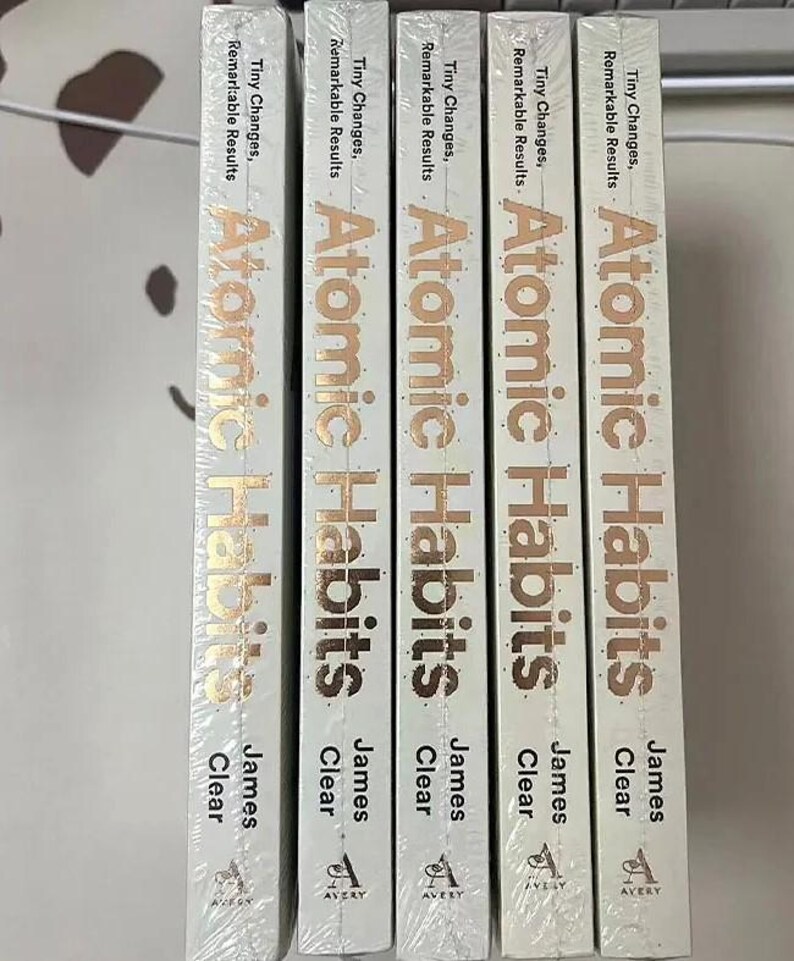 Peut inclure: Cinq exemplaires du livre "Atomic Habits" de James Clear sont pr&eacute;sent&eacute;s. Les dos des livres sont blancs avec des lettres dor&eacute;es. Le texte comprend "Tiny Changes, Remarkable Results" et le nom de l'auteur. Le logo Avery est en bas.