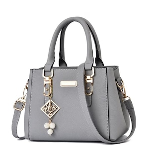 May include: A gray handbag with a top handle and a detachable shoulder strap. The bag features gold-colored hardware, including clasps and a rectangular accent. A decorative charm with a diamond-shaped pendant and white pearl-like beads hangs from the handle.