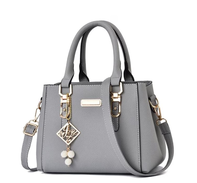 May include: A gray handbag with a top handle and a detachable shoulder strap. The bag features gold-colored hardware, including clasps and a rectangular accent. A decorative charm with a diamond-shaped pendant and white pearl-like beads hangs from the handle.