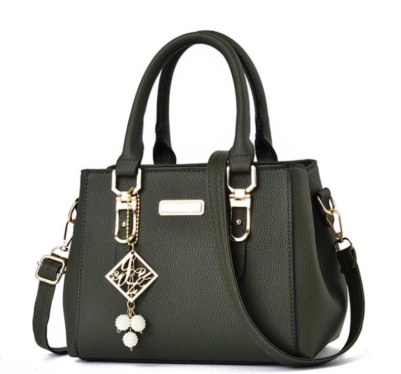 May include: Dark green handbag with top handles and a detachable shoulder strap. The bag features gold-colored hardware, including clasps and a decorative charm with white pearl accents. The bag has a textured surface and a rectangular shape.