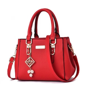 May include: A red handbag with a top handle and a detachable shoulder strap. The bag features gold-colored hardware, including clasps and a rectangular nameplate. A decorative charm with a geometric design and white beads hangs from the handle.