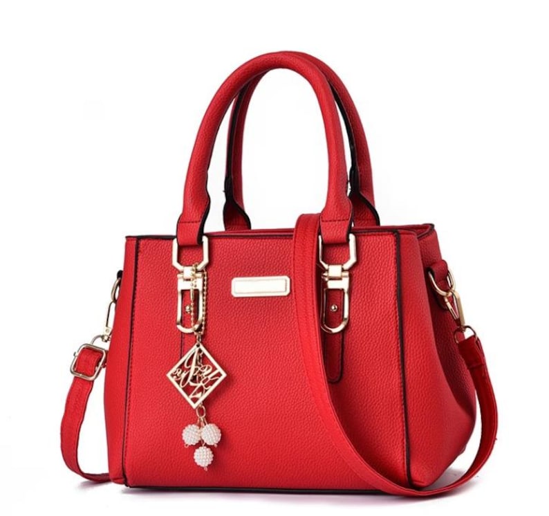 May include: A red handbag with a top handle and a detachable shoulder strap. The bag features gold-colored hardware, including clasps and a rectangular nameplate. A decorative charm with a geometric design and white beads hangs from the handle.