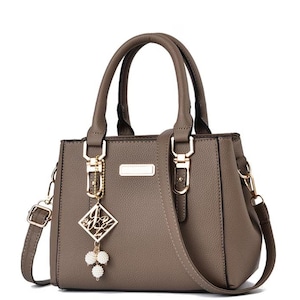 May include: A taupe-colored handbag with a top handle and a detachable shoulder strap. The bag features gold-colored hardware, including clasps and a decorative charm with a diamond-shaped pendant and pearl accents. The bag has a structured design.