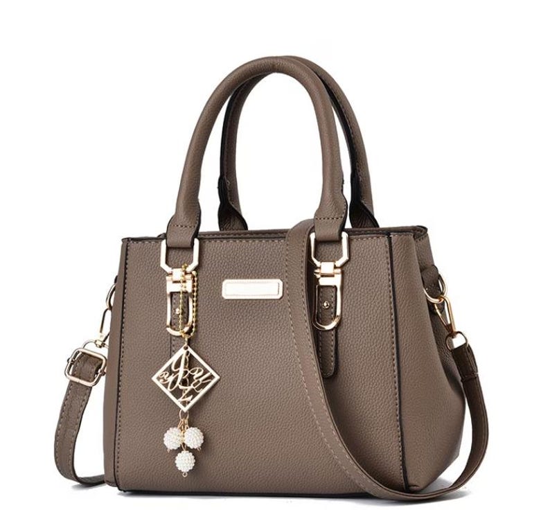 May include: A taupe-colored handbag with a top handle and a detachable shoulder strap. The bag features gold-colored hardware, including clasps and a decorative charm with a diamond-shaped pendant and pearl accents. The bag has a structured design.