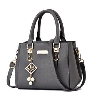 May include: Gray handbag with top handles and a detachable shoulder strap. The bag features gold-colored hardware, including clasps and a decorative charm with white pearl-like beads. The bag has a rectangular shape and a textured surface.