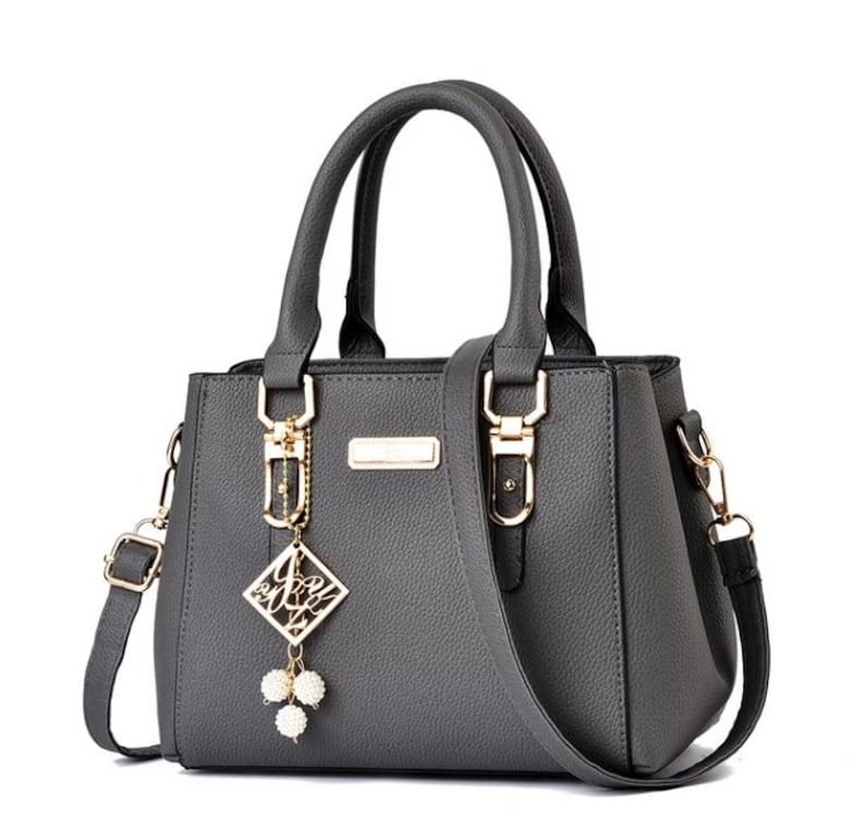 May include: Gray handbag with top handles and a detachable shoulder strap. The bag features gold-colored hardware, including clasps and a decorative charm with white pearl-like beads. The bag has a rectangular shape and a textured surface.