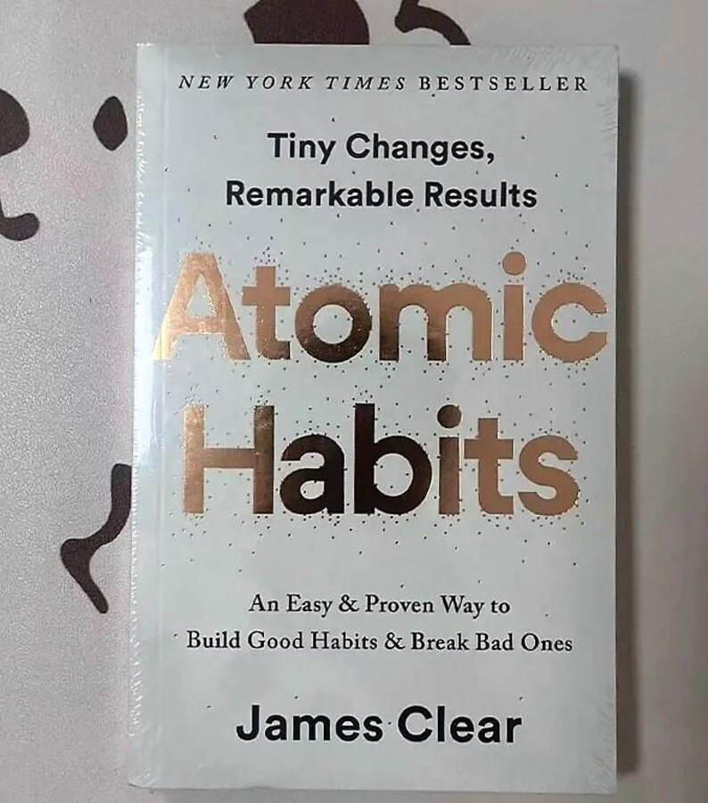 Peut inclure: Livre blanc intitul&eacute; "Atomic Habits" avec des lettres dor&eacute;es. Le livre est un best-seller du New York Times et comprend le nom de l'auteur, James Clear. La couverture indique &eacute;galement "Tiny Changes, Remarkable Results".