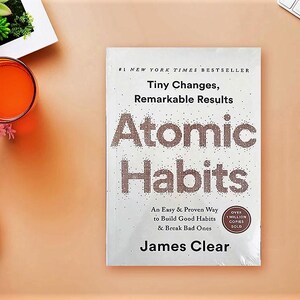 Peut inclure: Un exemplaire de "Atomic Habits" de James Clear, un best-seller du New York Times, est pos&eacute; sur une surface couleur p&ecirc;che. Le titre du livre est bien en &eacute;vidence, avec le slogan "Tiny Changes, Remarkable Results."
