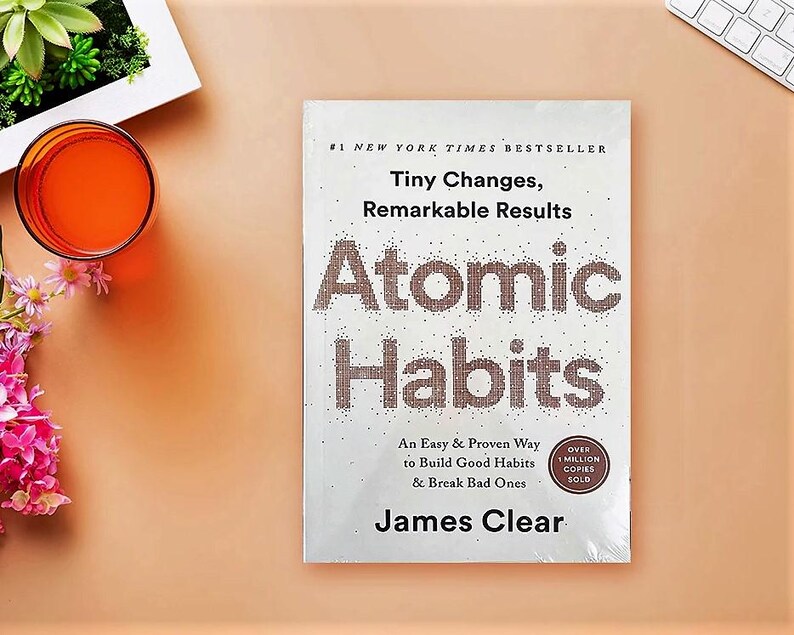Peut inclure: Un exemplaire de "Atomic Habits" de James Clear, un best-seller du New York Times, est pos&eacute; sur une surface couleur p&ecirc;che. Le titre du livre est bien en &eacute;vidence, avec le slogan "Tiny Changes, Remarkable Results."