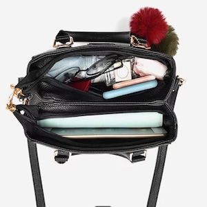 May include: Black leather handbag with gold-tone hardware, open to reveal contents. Includes glasses, a tablet, and other personal items. A red and green pom-pom charm hangs from the handle. The bag has a top handle and a shoulder strap.