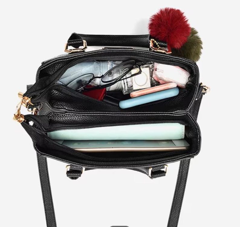 May include: Black leather handbag with gold-tone hardware, open to reveal contents. Includes glasses, a tablet, and other personal items. A red and green pom-pom charm hangs from the handle. The bag has a top handle and a shoulder strap.