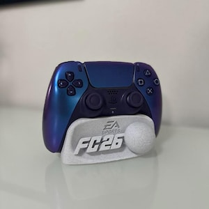 Op de afbeelding: Een blauw-paarse PlayStation 5-controller op een witte standaard. De standaard toont "EA SPORTS FC26" en een voetbal. De controller heeft een donkerpaars midden.