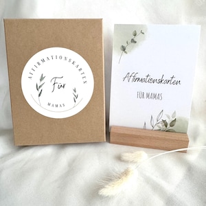 May include: A set of affirmation cards for mothers, displayed with a wooden card holder and a kraft paper box. The cards feature botanical illustrations and the text "Affirmationskarten für Mamas."